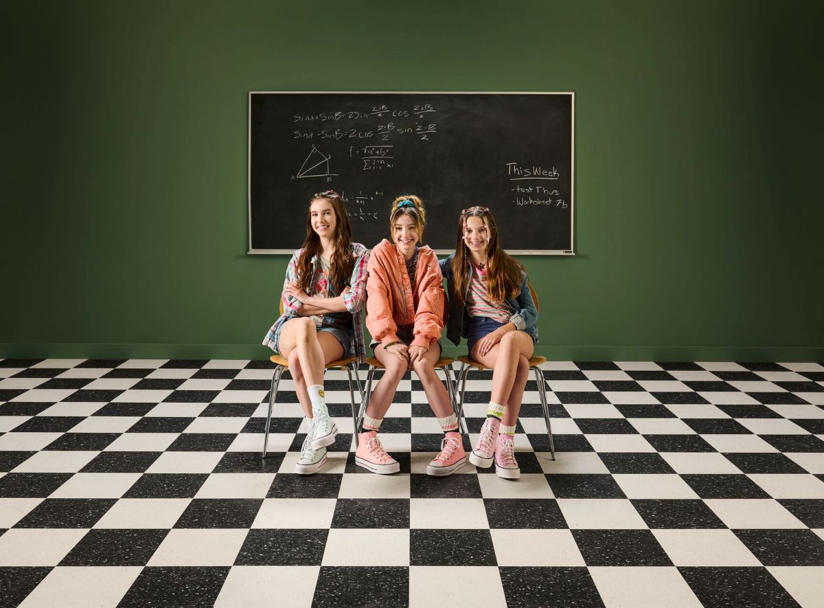 Three young female students sitting on stools in front of a chalkboard; checkered flooring pattern showing unique K-12 design